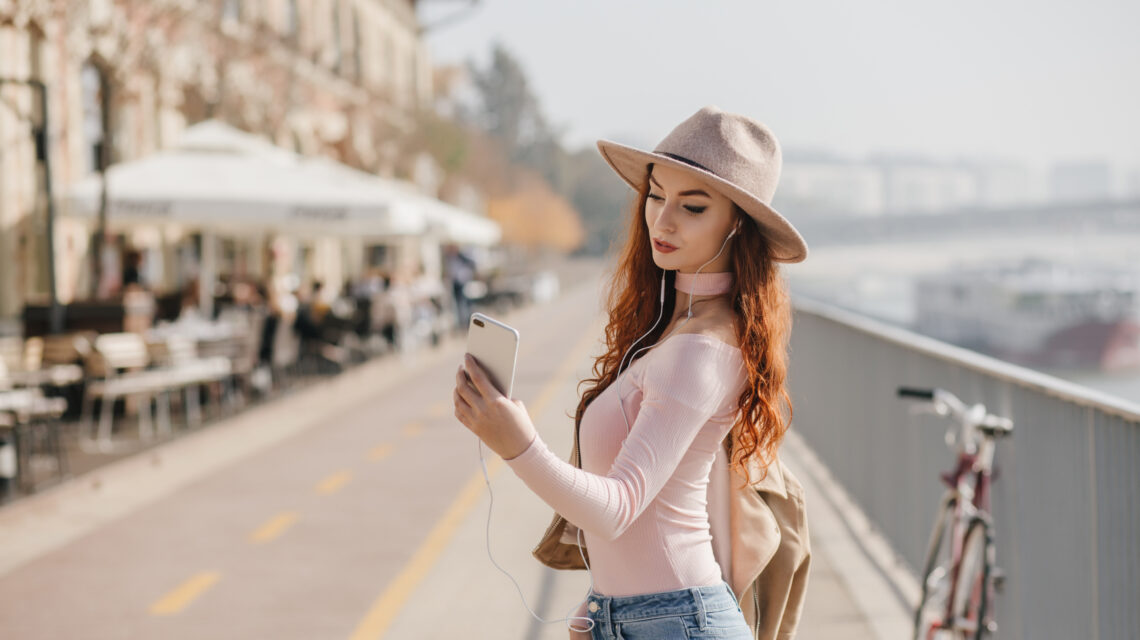 Serious young woman with wavy ginger hair making selfie. Outdoor photo of slim girl in pink shirt taking picture of herself at embankment..