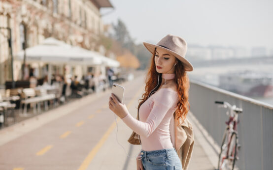 Serious young woman with wavy ginger hair making selfie. Outdoor photo of slim girl in pink shirt taking picture of herself at embankment..